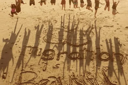 360 students jump above the text "Migration is Beautiful" carved in the sand of a beach