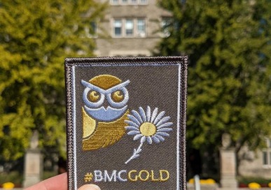 A hand holding an iron-on patch in front of Pem Arch. The patch is gray and features an owl, a daisy, and the words "#BMCGOLD."