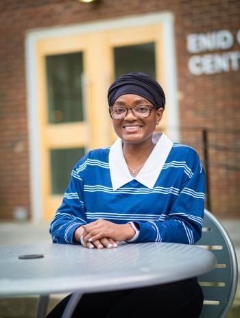 Saeina Charles smiling and seated at a table in front of the Enid Cook Center