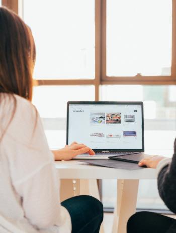 Two persons look at content displayed in a laptop. Their back is to the camera.