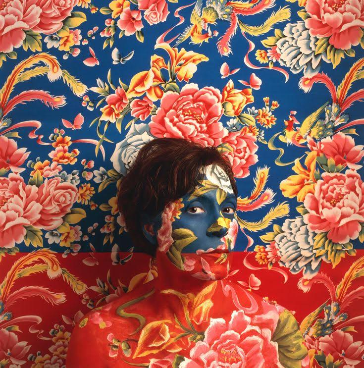 Self Portrait of Artist wearing blue and red floral make-up as camouflage with wallpaper behind her