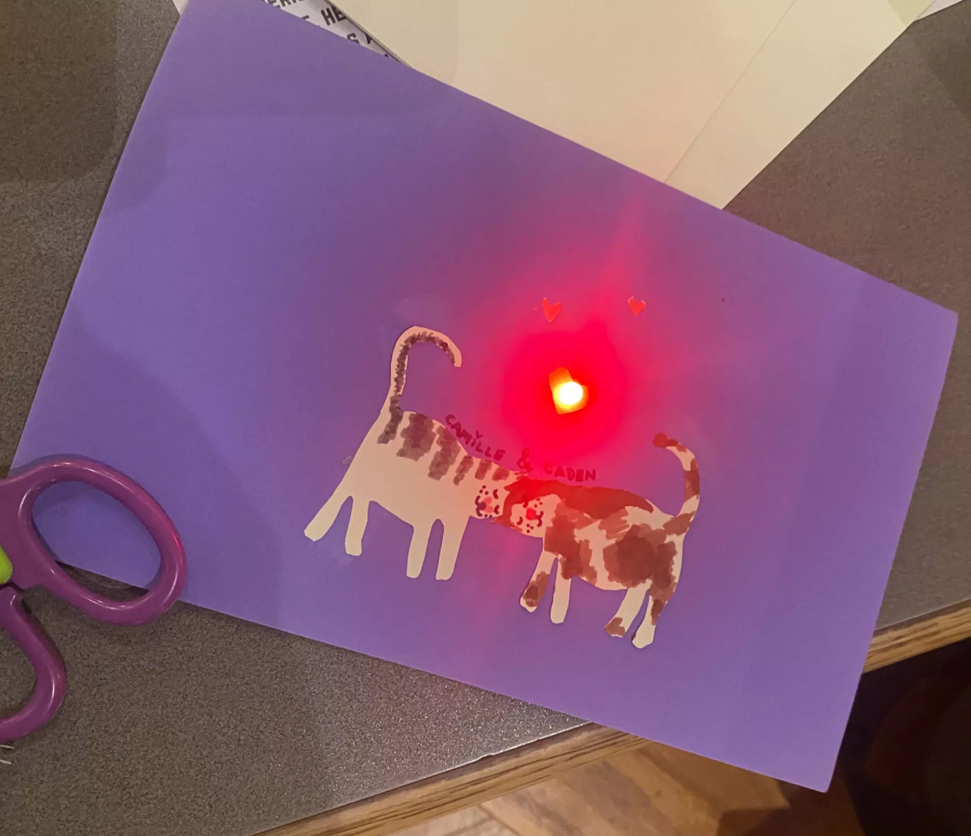Valentine's Day card made during student workshop. The card is purple and reads, "Camille & Caden." There are then two cartoon cats underneath the text. They are rubbing heads and a red light is shining where their heads meet.