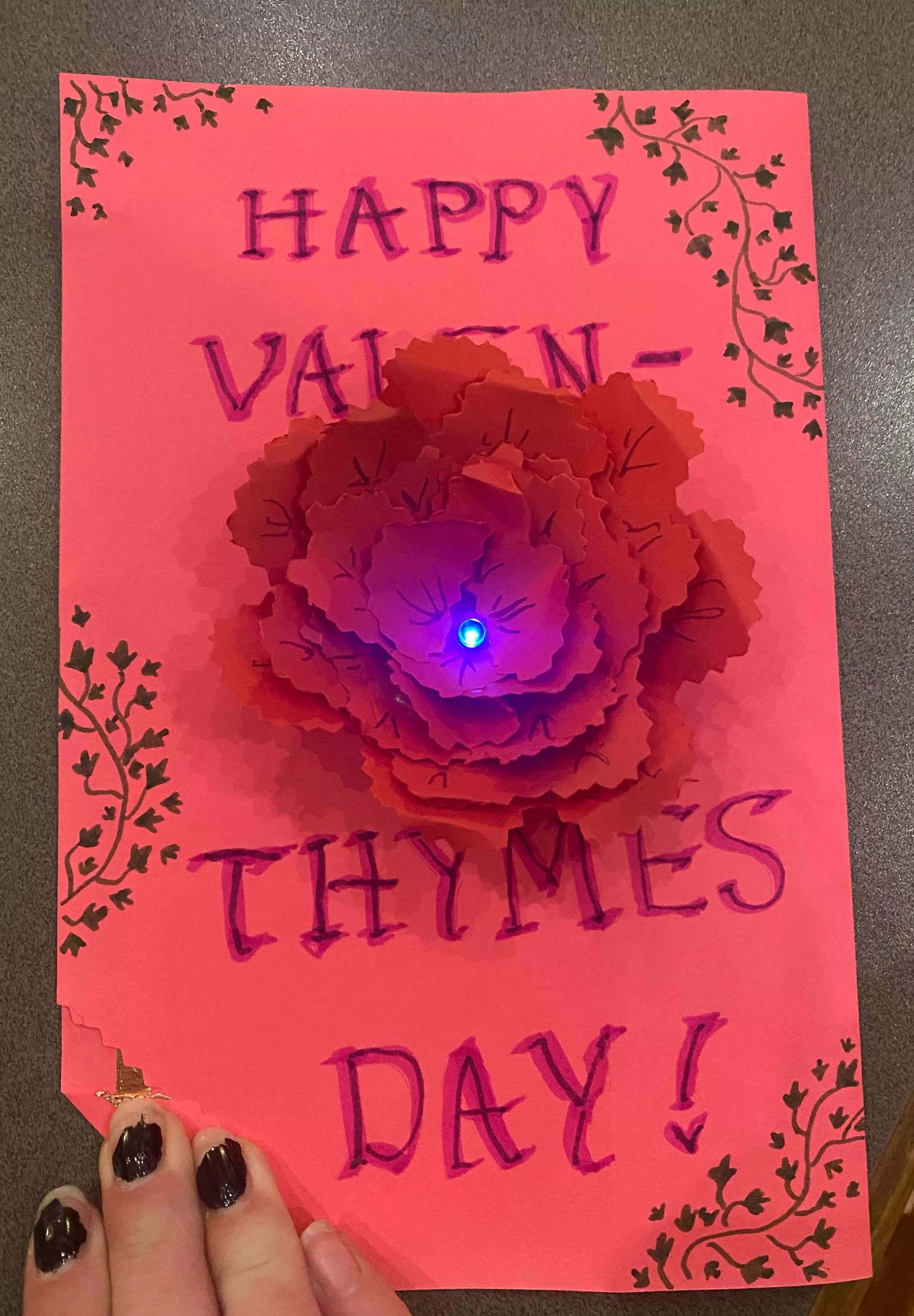 Pink Valentine's Day card made during student workshop. There's an origami rose in the middle of the card with a blue light in the middle of it. The card reads, "Happy Valentines Day!"
