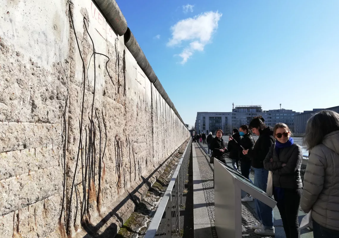 A stone wall with line drawings of figures on the left side of the frame; a row of 6 students and faculty on the right side of the frame observe and read a plaque about the art under a blue sky.