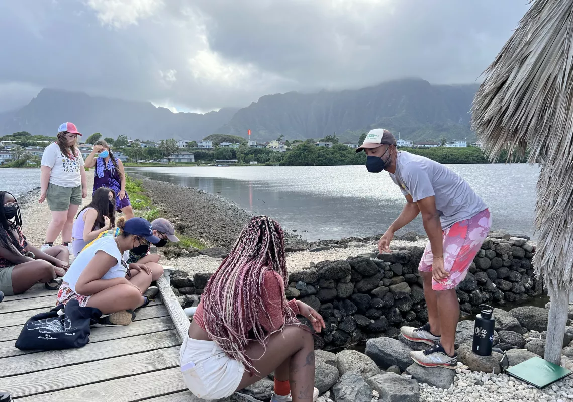 Six students cluster on a wooden platform with volcanic mountains, voluminous grey clouds, and a calm lake in the background. A student and a facilitator converse in the foreground, standing on large rocks near a retaining wall.