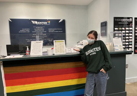 Elana standing in front of a desk with a Rainbow flag