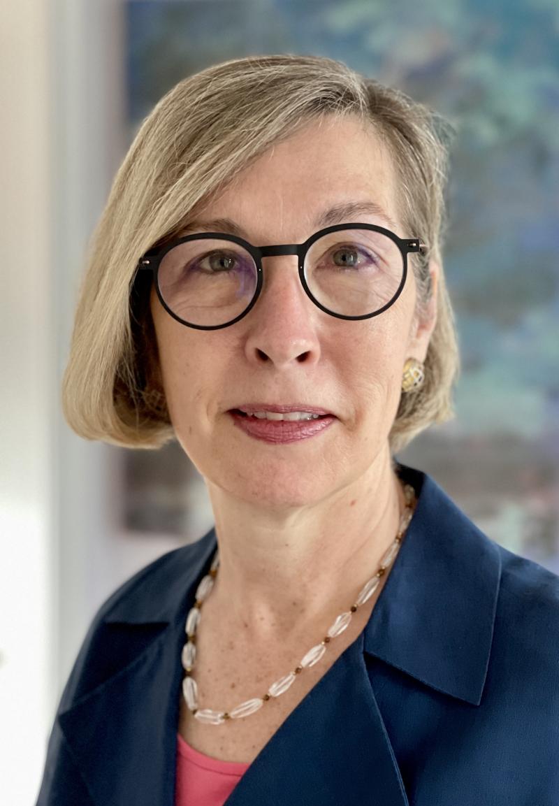 A woman with jaw length blond hair looks at the camera. She is wearing round glasses, a navy blazer, and a pink shirt.